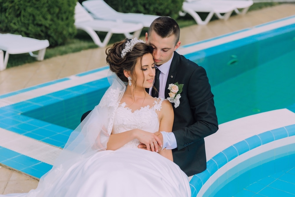 A bride and groom are sitting on a poolside, with the bride wearing a white dress and the groom wearing a suit