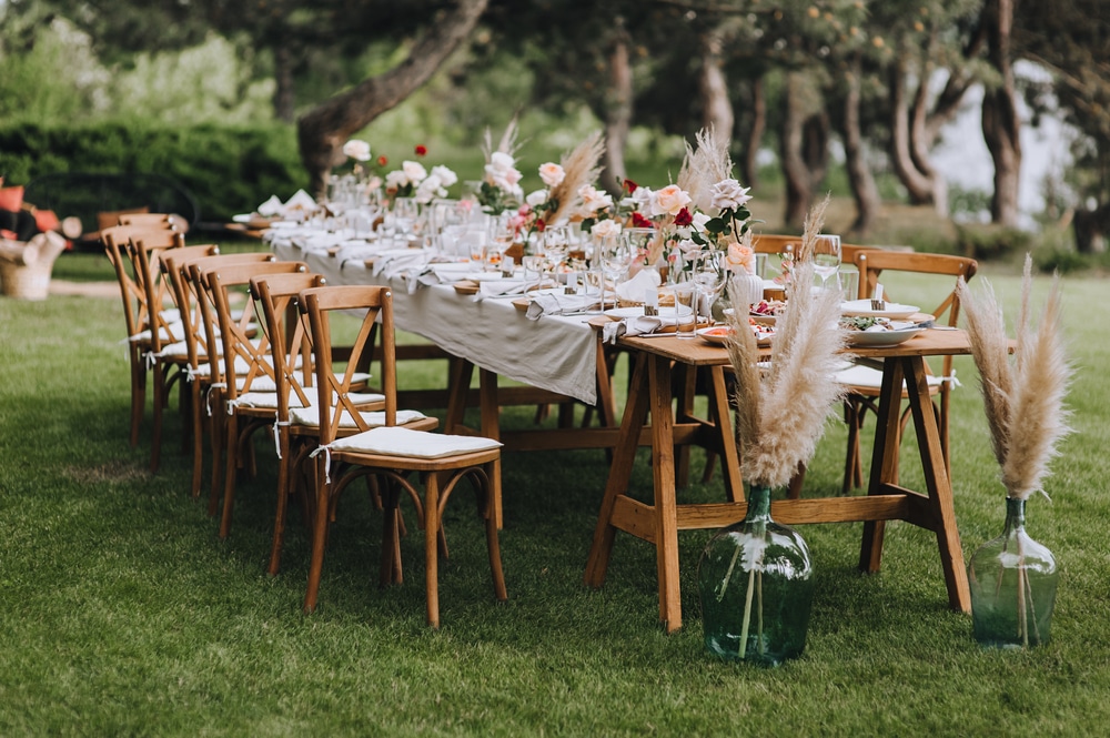 A large, long, decorated, wooden table and chairs, covered with a white tablecloth with dishes, flowers, candles, stands on the green grass in the park, in the forest in nature.