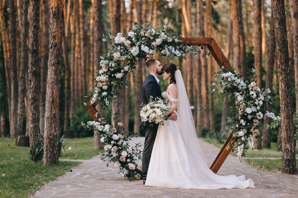 Stylish groom in a suit and a cute brunette bride in a white dress in the forest near a wedding wooden arch decorated with flowers.