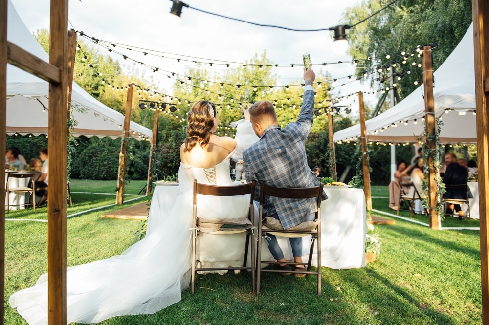 Young bride and groom sit at the table in decorated backyard and greet their guests