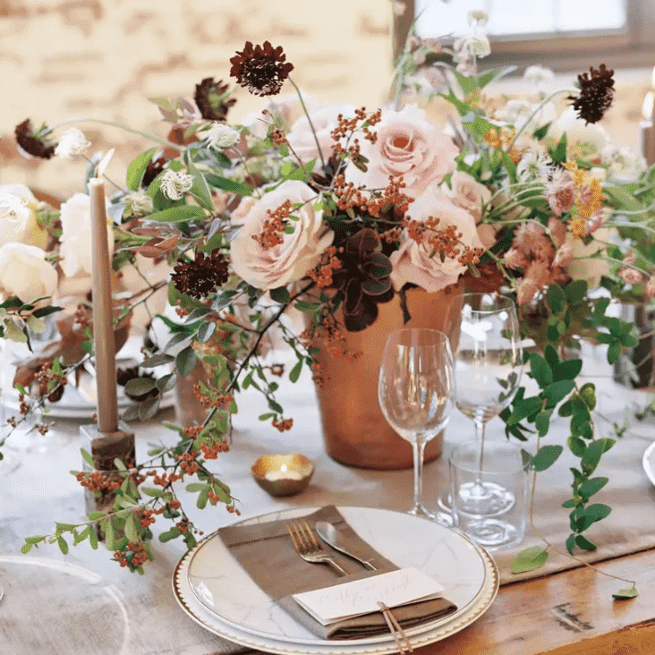 terracotta and sage green flowers in a vase on a table along with glass table plates and candles